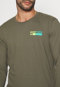 Man in een olijfgroene langemouwen shirt met neon "Project Rock" tekst en een stierlogo op de linkerborst.