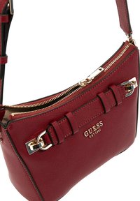 Sac à main Guess en cuir rouge avec fermeture éclair dorée, boucle décorative et logo "GUESS EST 1981" à l'avant.