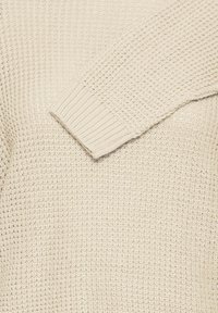 Suéter de punto beige con mangas largas, que presenta una textura gruesa de tejido en waffle y puños acanalados.