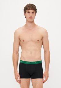 Schwarze Boxer-Shorts mit einem grünen Bund, auf dem das Logo "TOMMY HILFIGER" prangt. Der Stoff wirkt weich und glatt und hat ein enganliegendes Design.