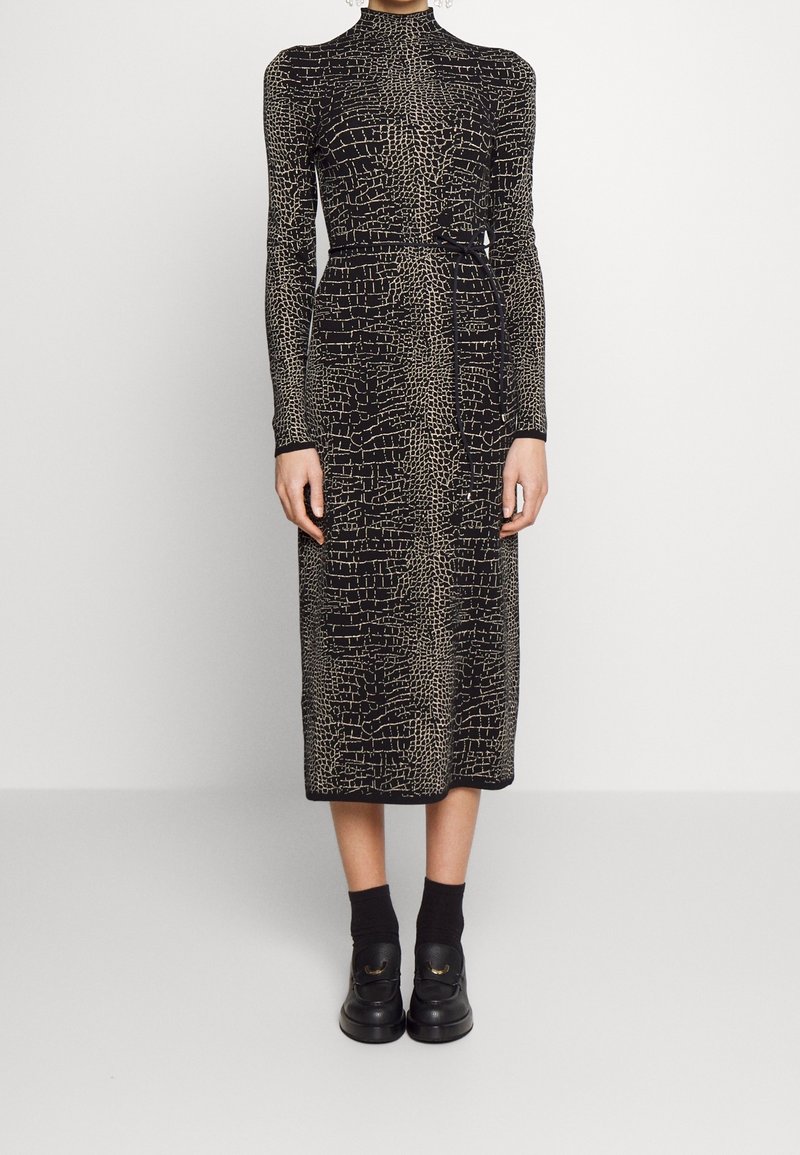 Femme portant une robe noire à manches longues et col montant avec un motif abstrait beige et une fine ceinture, associée à des chaussettes noires et des mocassins.