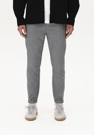 Man in grijze slim-fit geruite broek, witte sokken, lichtgrijze sneakers, een wit shirt en een zwarte knopenjas, staand tegen een witte achtergrond.