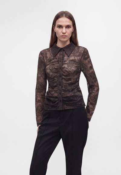 Femme aux cheveux lisses portant une chemise noire en dentelle ajustée à manches longues, boutonnée, et un pantalon noir sur un fond clair uni.