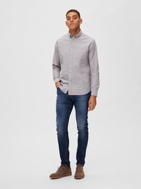 Camisa de botones gris con cuello y bolsillo, combinada con vaqueros azul oscuro y zapatos de ante marrones, de pie frente a un fondo liso.