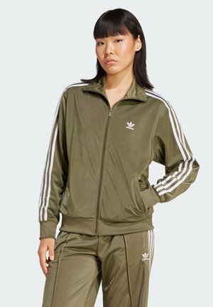 Donna che indossa un tracksuit Adidas verde oliva con strisce bianche, in piedi con una mano in tasca su uno sfondo chiaro e neutro.