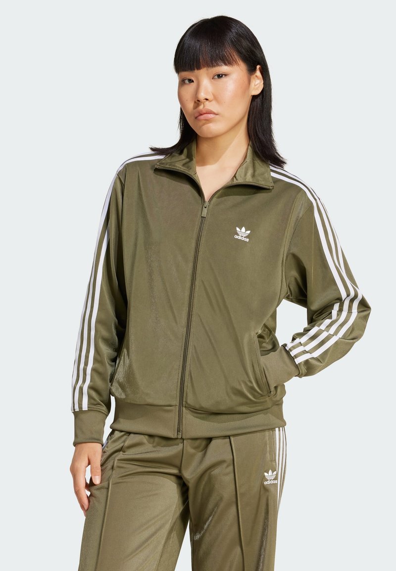 Žena u maslinasto zelenom Adidas trening odijelu s bijelim prugama, stoji s jednom rukom u džepu na jednostavnoj svijetloj pozadini.
