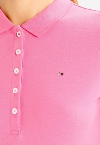 Różowa koszulka polo o dzianinowej strukturze, z trzema guzikami i małym haftowanym logo w czerwono-biało-niebieskich kolorach na piersi.