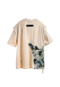 Camisa de manga corta color beige con una etiqueta rectangular negra en la parte superior de la espalda y un gran parche de tela texturizada en tonos azules y verdes en la parte inferior derecha.