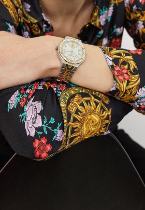 Håndled med sølv- og guldkolouret ur med rhinsten båret over en foldet sort ærme med blomstret og gyldent solmønster.