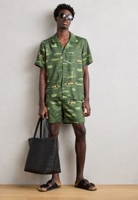 Grøn kortærmet skjorte og matchende shorts med mønster af bygninger og køretøjer. En sort tote bag og sandaler fuldender looket.
