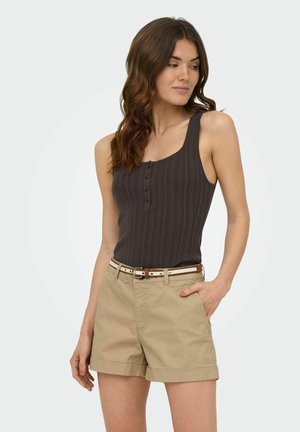 Femme aux longs cheveux ondulés portant un haut sans manches côtelé gris foncé et un short beige avec une ceinture blanche et marron, debout avec une main dans la poche.