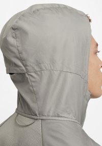 Perfil lateral de una persona que lleva una chaqueta con capucha de color gris claro, fabricada con tejido texturizado, sobre un fondo blanco.