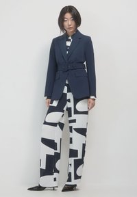 Blazer navy con cintura, abbinato a pantaloni a gamba larga con motivo bianco e nero. Tessuto liscio, vestibilità strutturata, rever affilati e tacchi a punta.