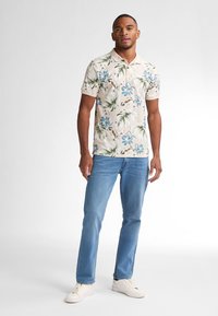Polo a maniche corte di colore beige chiaro con motivo floreale blu e foglie verdi. Abbinato a jeans azzurri e sneaker bianche.