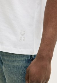 Biała bawełniana koszulka z nadrukiem logo z boku "ID EST: 2007" w jasnoszarym kolorze. Materiał wydaje się być miękki o gładkiej teksturze.