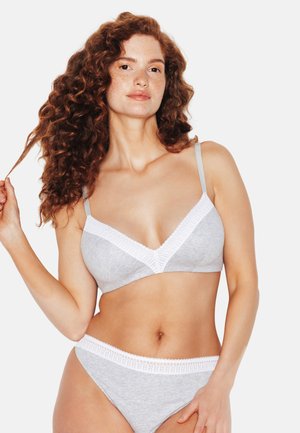 Donna con capelli rossi e ricci che indossa biancheria intima grigia con bordatura in pizzo bianco, tenendo una ciocca di capelli con un’espressione neutra.