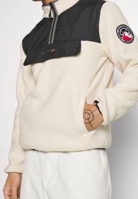Fleece-pullovern i krämfärgad med en svart nylonbröstpanel, dragkedja vid halsen, ficka och logotypmärke, med kontrasterande texturer och färger.