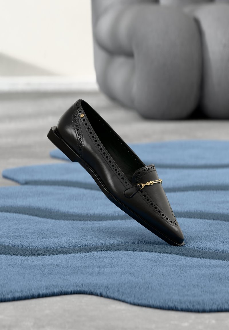 Mocassin en cuir noir avec détails perforés et boucle dorée posé sur un tapis bleu texturé à formes abstraites.