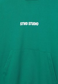Grøn hættetrøje med en lomme foran og hvid tekst "STWD STUDIO" centreret over lommen.