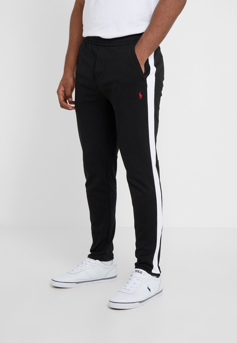 Man som bär svarta träningsbyxor med vita sidostreck och röd logotyp, i kombination med vita sneakers med svarta logotyper.