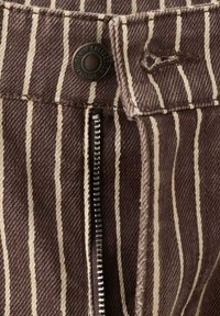 Hnedá rifľovina s bielymi vertikálnymi pruhmi, kovový zips a gombík s nápisom "Original Denim Collection" na páse odevu.