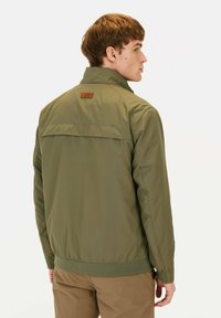 Olivgrüne Nylonjacke mit hohem Kragen, elastischen Bündchen und einem Rückeneinsatz. Mit einem braunen Lederpatch im oberen Rückenbereich.