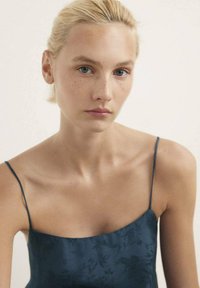 Camisola in satin blu scuro con spalline sottili, caratterizzata da delicati motivi floreali e una texture liscia, indossata da un modello con un'espressione neutra.