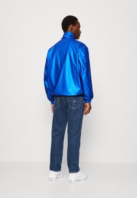 Blå, skinnende jakke med høy krage og lange ermer, kombinert med mørk denimjeans og hvite joggesko, sett bakfra.