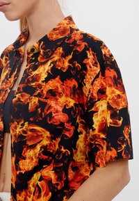 Kortärmad skjorta med svart bakgrund som har ett livfullt flamamönster i nyanser av orange och gult; inkluderar en bröstficka och knäppning.