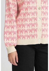 Cardigan beige clair avec un motif répétitif de nœuds roses, des poignets et un ourlet côtelés, et de grands boutons clairs sur le devant.