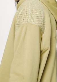 Beige hoodie showing close-up of shoulder with perforated mesh fabric detail and part of hood visible.