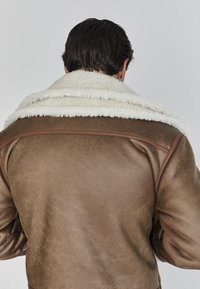 Chaqueta de cuero marrón con un suave cuello de borreguito blanco. La textura parece desgastada, mostrando un acabado rústico y un diseño de cuello contrastante.