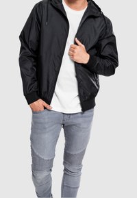 Black athletic zip-up jacket with a hood, ribbed cuffs, and smooth texture; worn over a white T-shirt and paired with light grey jeans.