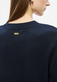 Μπλε πλεκτό πουλόβερ με στρογγυλή λαιμόκοψη. Διαθέτει μικρή χρυσή ετικέτα με λογότυπο στο πίσω μέρος. Λείο υλικό; μεσαίο πάχος.