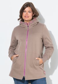 Donna con capelli castani che indossa una felpa taupe con zip rosa e dettagli rosa sul colletto, con le mani in tasca, in piedi su uno sfondo bianco.