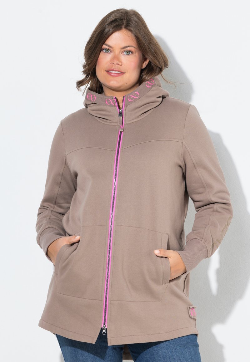 Donna con capelli castani che indossa una felpa taupe con zip rosa e dettagli rosa sul colletto, con le mani in tasca, in piedi su uno sfondo bianco.