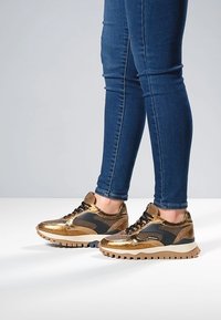Metallic gold and black sneakers with textured surfaces, lace-up design, and contrasting sole patterns, paired with fitted blue jeans.