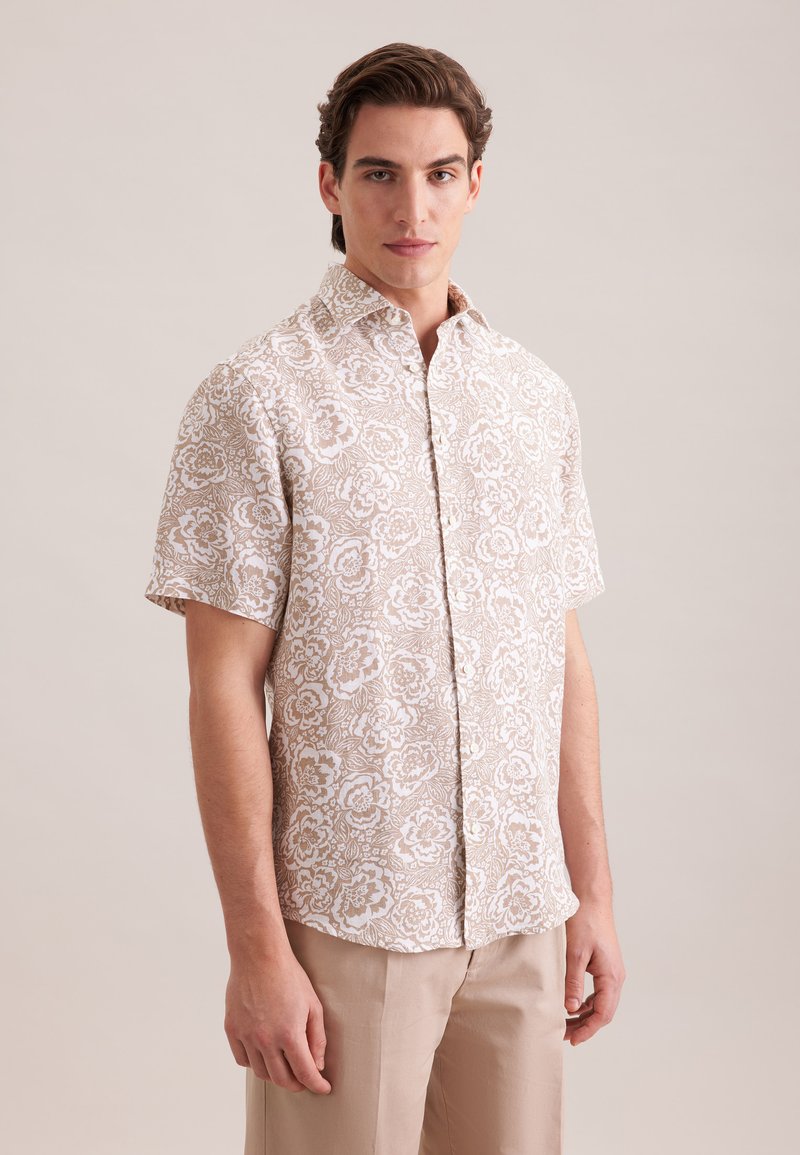 Homme portant une chemise à manches courtes à boutons beige et blanche à motifs floraux, avec un pantalon beige, debout devant un fond uni clair.