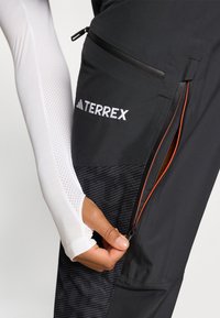 Zwarte Terrex-broek met ritszakken en oranje accenten. Close-up van een hand die naar de zak reikt, waarbij het gladde materiaal en de gestructureerde stof zichtbaar zijn.