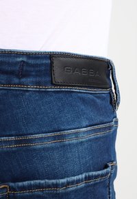 Mörkblå denimjeans med en lädermärke som har texten "GABBA" på midjan, med synliga sömmar och två bakfickor.