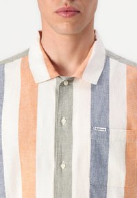 Camisa de botão com riscas verticais em laranja, branco, cinza e azul. Inclui um bolso no peito e uma pequena etiqueta da marca.