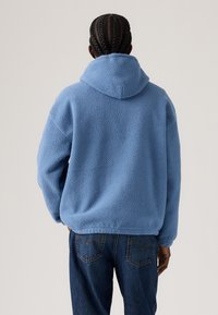 Sweat à capuche en polaire bleue avec une surface texturée et douce. Comprend une grande capuche et une coupe décontractée, associé à un jean en denim noir.