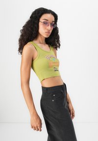 Young woman with long curly hair wears green cropped tank top, purple-tinted glasses, and high-waisted black skirt, posing sideways.