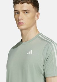 T-shirt de sport vert clair en tissu respirant. Présente des rayures blanches sur les épaules et un petit logo blanc sur la poitrine.