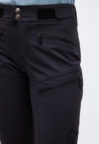 Nærbilde av svarte bukser med to glidelåslommer på låret og hoften, dobbel knappestenging og justerbar velkrobelte i midjen.