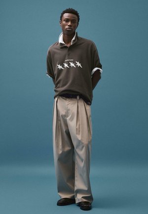 Mladenič stoji pred modrim ozadjem, oblečen v temno rjavo polo majico z vzorcem zvezd in bež široke hlače, roki ima za hrbtom.