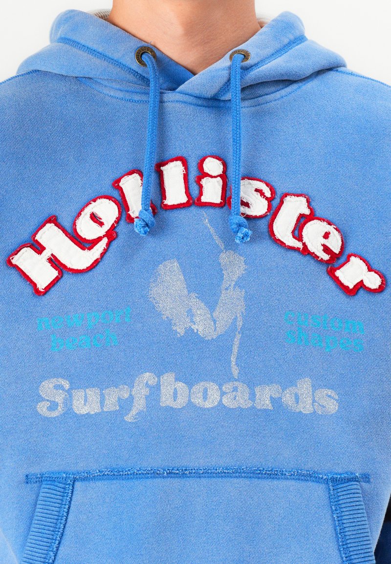 Sudadera azul claro con un bolsillo canguro. Presenta texto bordado en rojo y blanco en un estilo audaz y acentos impresos en plateado. Cuerda y ojales de metal.