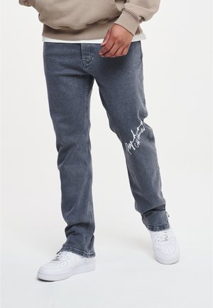Personne portant un jean noir délavé avec une écriture blanche sur la cuisse et des baskets blanches, sweat beige partiellement visible.