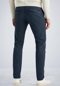 Chino in cotone blu navy con vestibilità slim, tasche posteriori e dettagli con bottone, abbinati a sneaker bianche. Texture liscia e linee pulite.