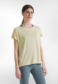 Lysegul kortærmet t-shirt med l�øs pasform. Blød tekstur, rund halsudskæring, lagdelt over en blå sports-bh, kombineret med teal farvede leggings.